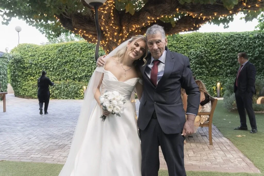 Novia con su ramo junto a un hombre de traje, bajo una pérgola con luces