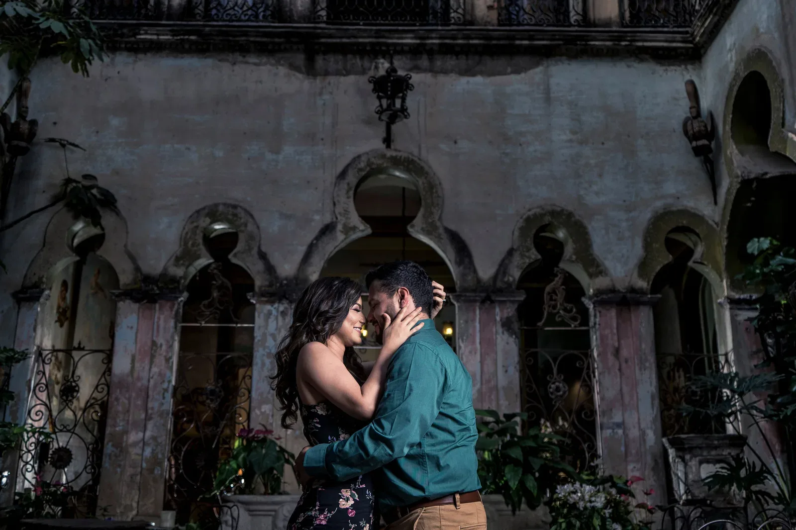 Pareja abrazada de noche en un patio con arcos, ella con vestido floral oscuro y él con camisa verde