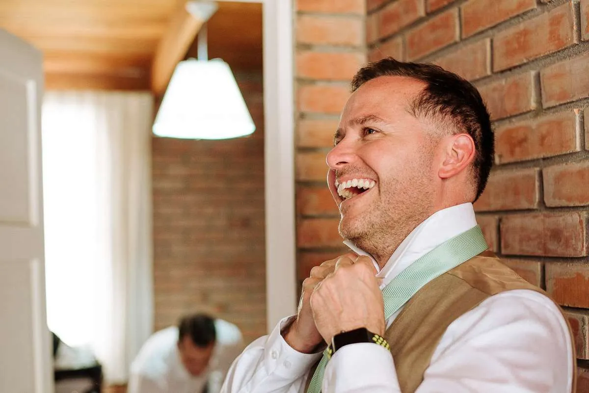 Groom laughing while adjusting his shirt in a wood-panelled room