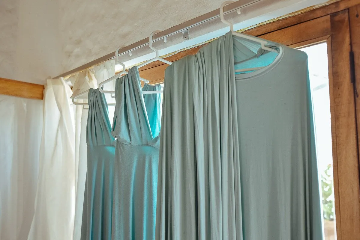 Three bridesmaid dresses in grey-green hanging from a rail beside a window with curtains