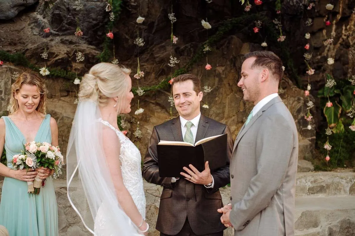 The couple before the officiant, with a rock wall and flowers behind
