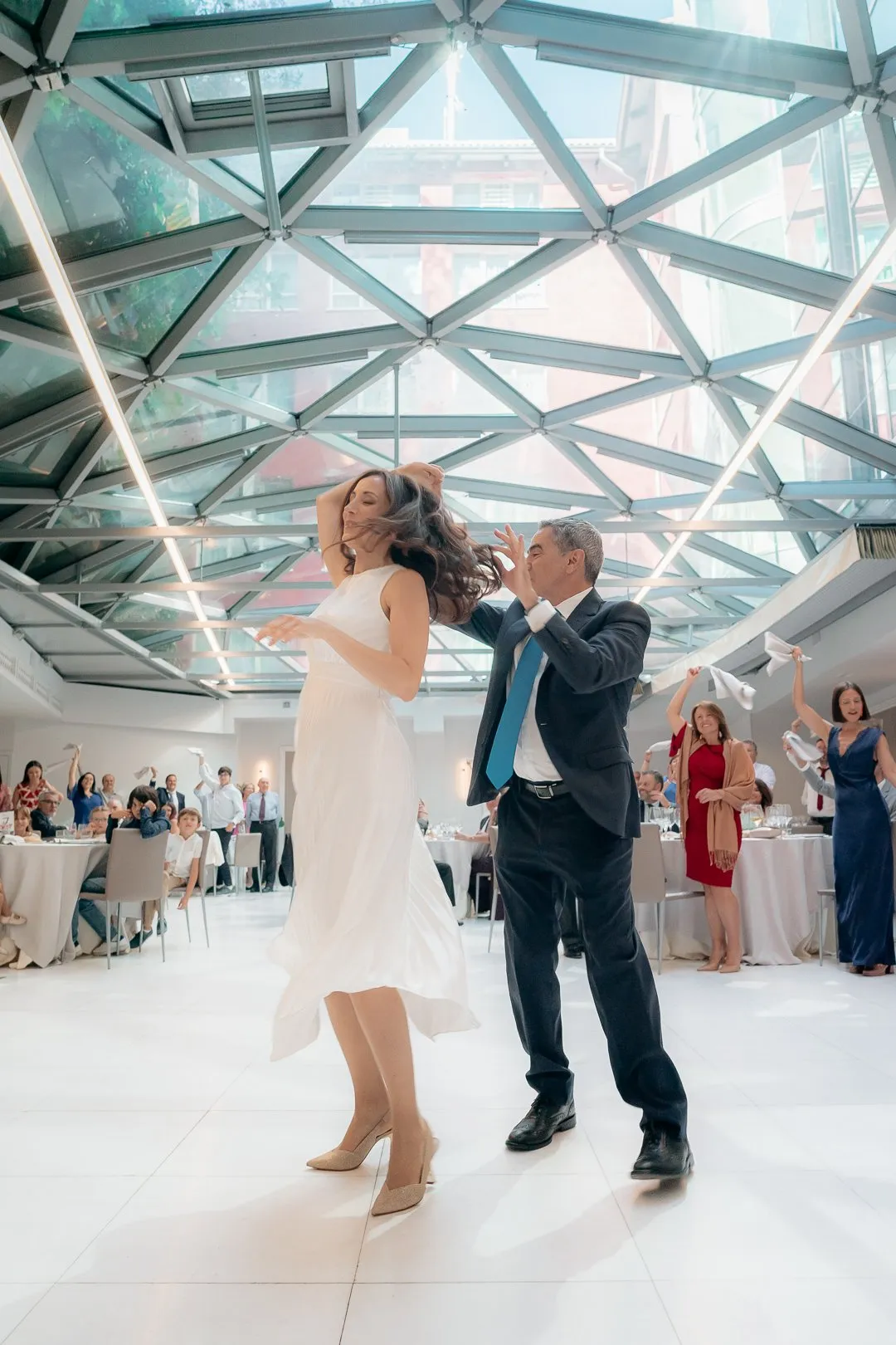 The couple dance beneath the glass roof of a hotel ballroom, with guests waving napkins behind them