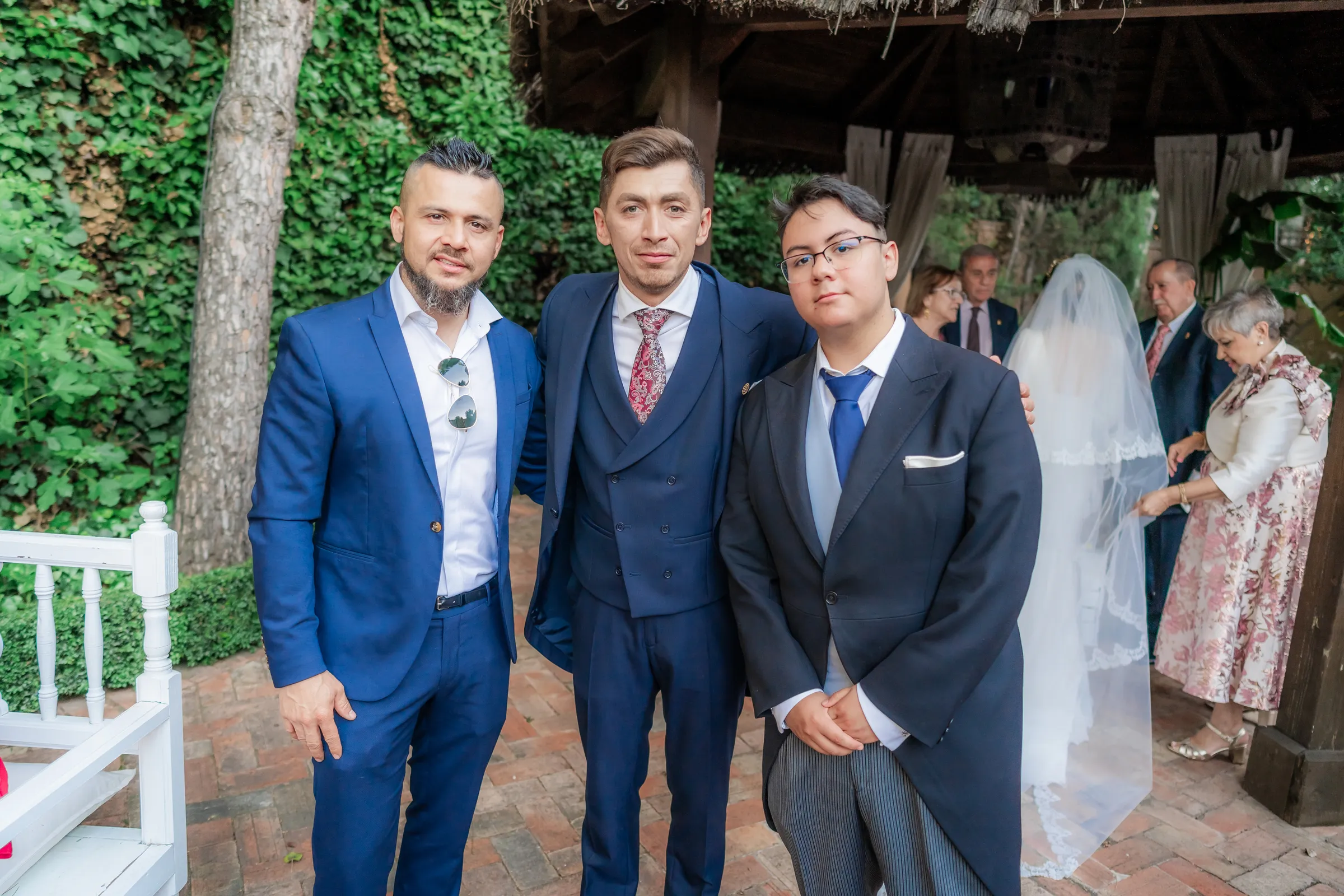 Tres amigos con traje azul en un jardín, con una novia al fondo