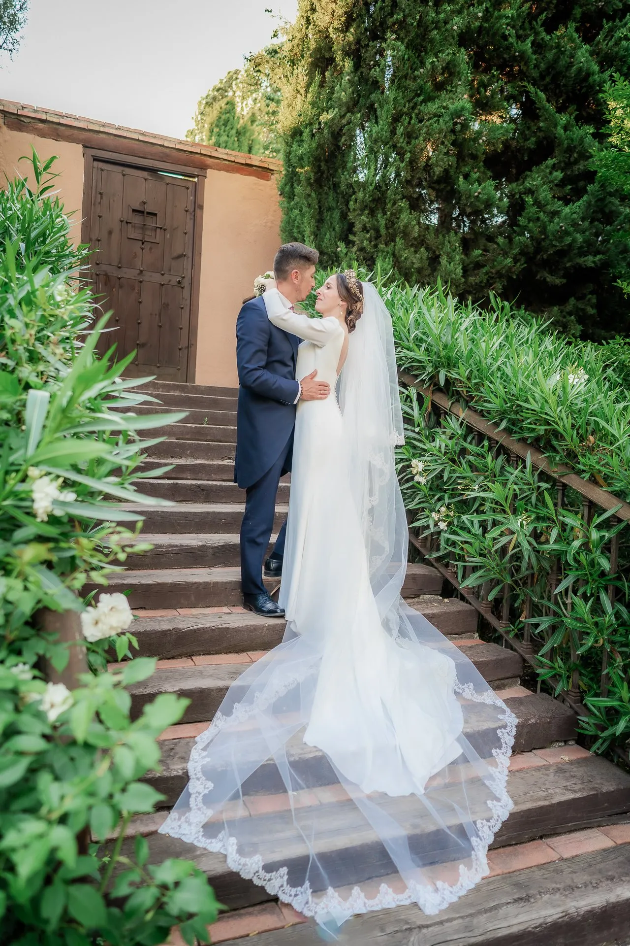 The couple embrace on a stone staircase among green shrubs, with the long veil spread over the steps