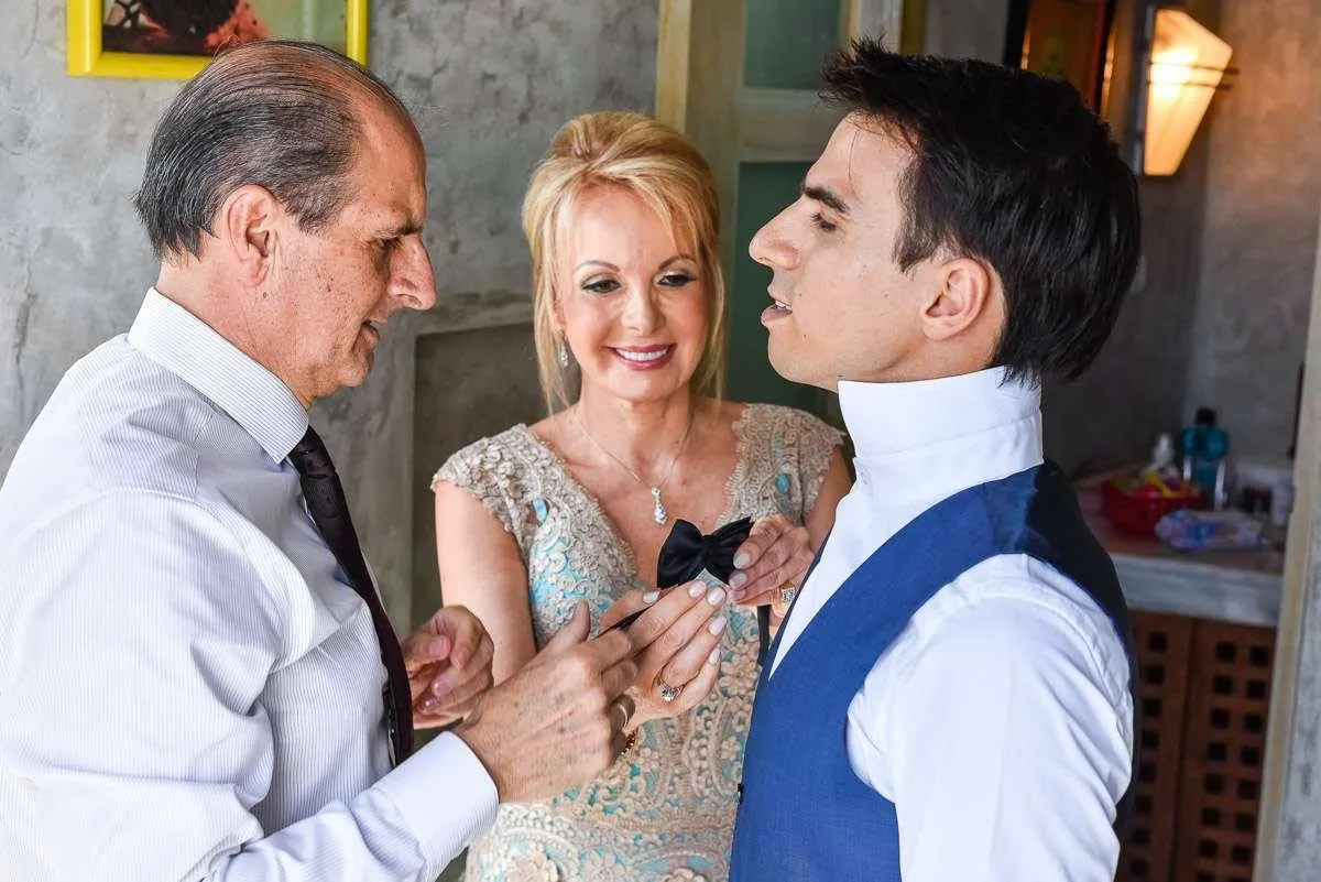 An older man and woman adjust the outfit of a young man in a blue waistcoat and bow tie, before the ceremony