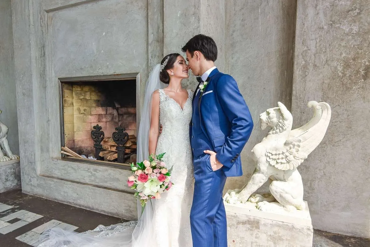 The bride and groom, alone beside a stone fireplace and a winged sculpture, look at each other very close