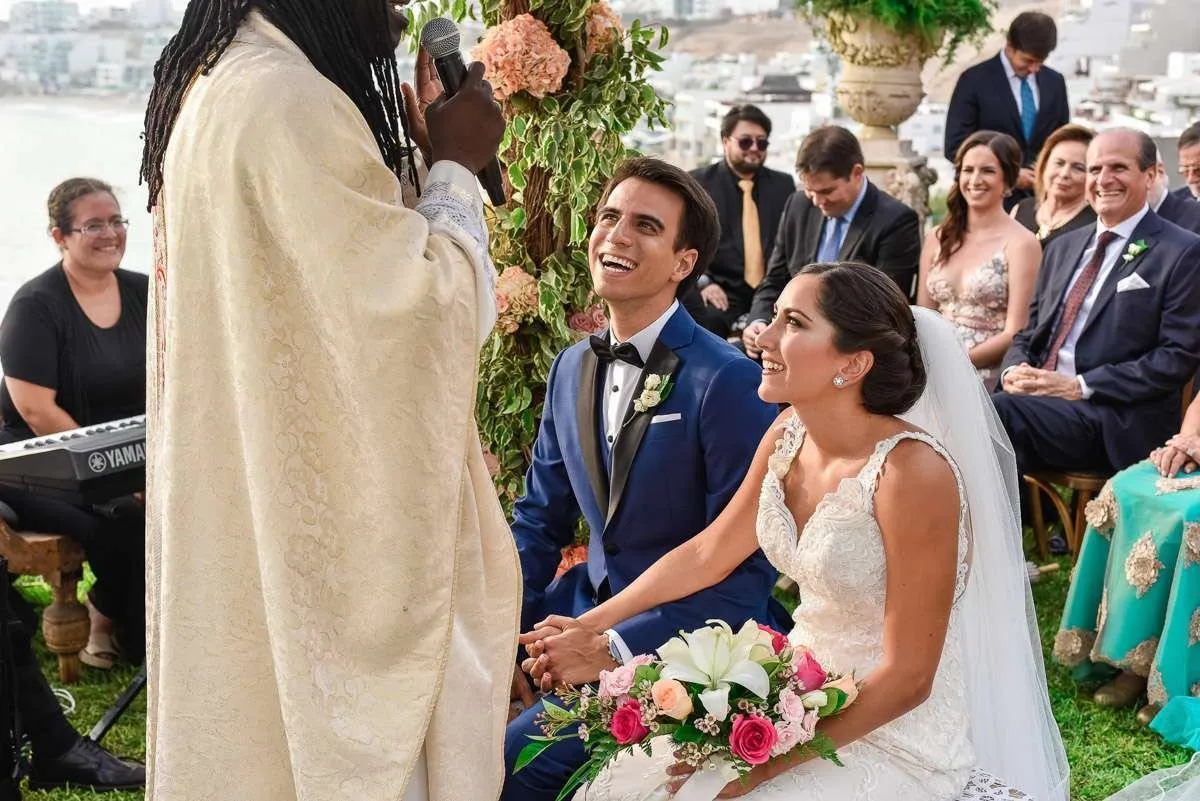The couple, seated and smiling, listen to the officiant, with guests behind them