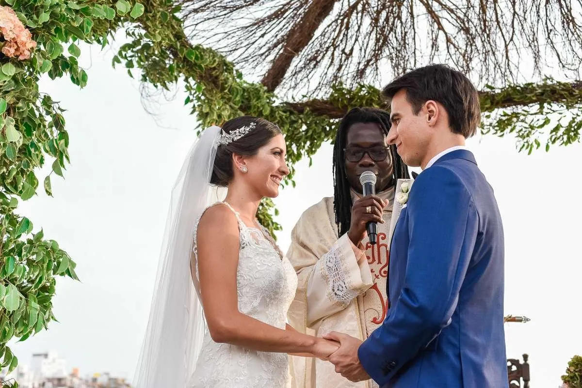 The couple exchange rings before the officiant, who speaks into a microphone under a bower of branches and flowers