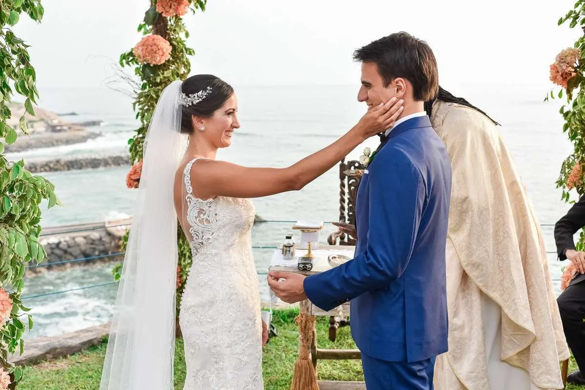 The bride touches the groom's cheek before an arch of flowers and branches, with the sea behind and the officiant to one side