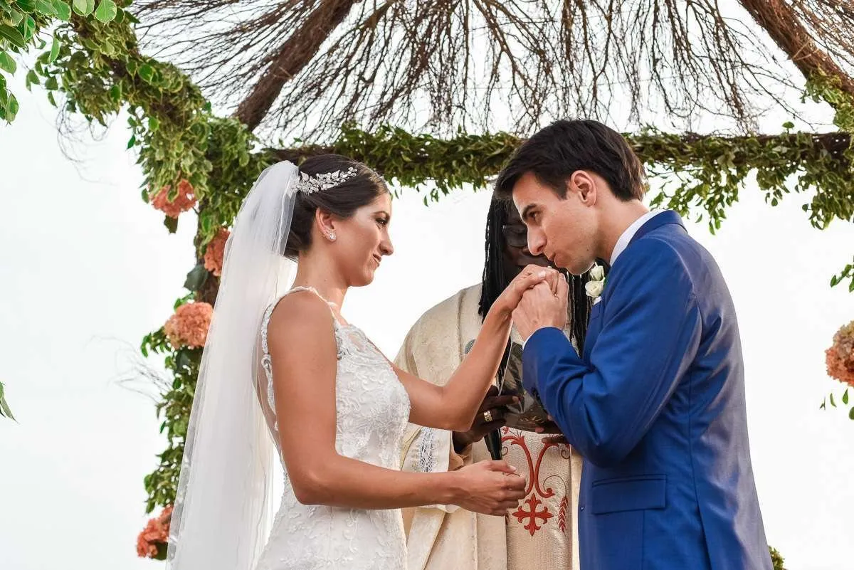 The couple, face to face and holding hands, before the officiant under a bower of branches and flowers