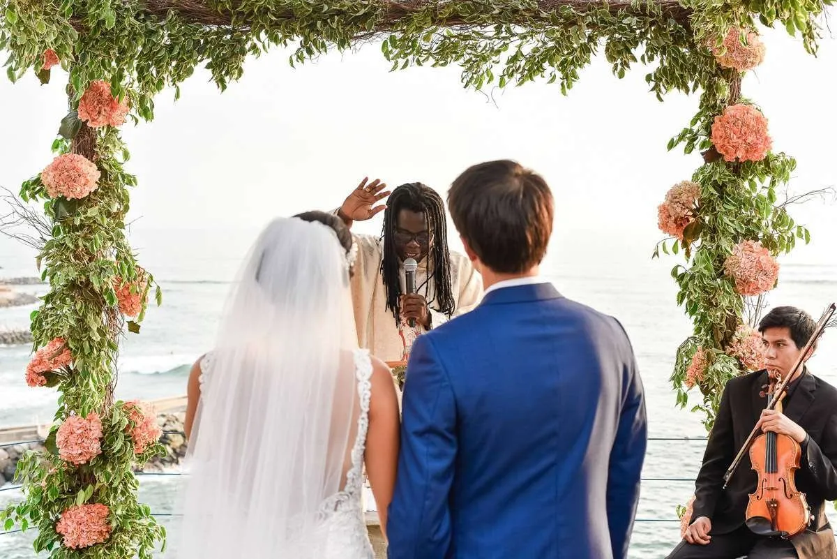 Couple embracing under a flower arch by the sea, with a violinist behind