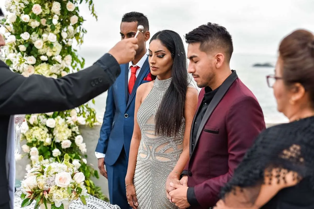 The officiant raises a hand to bless the couple beside an arch of white flowers, with the sea behind