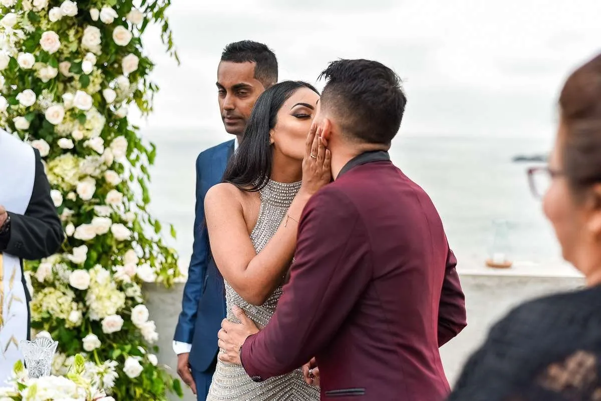 A guest kisses the bride on the cheek beside an arch of white flowers, with the sea behind