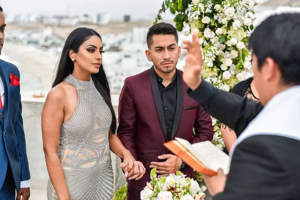 The couple hold hands as the officiant reads from an open book beside an arch of white flowers