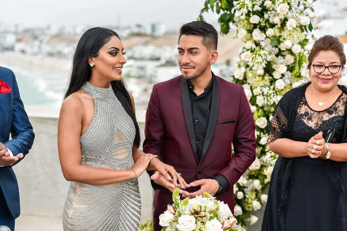 The couple, facing each other, join hands beside an arch of white flowers, with a guest at their side