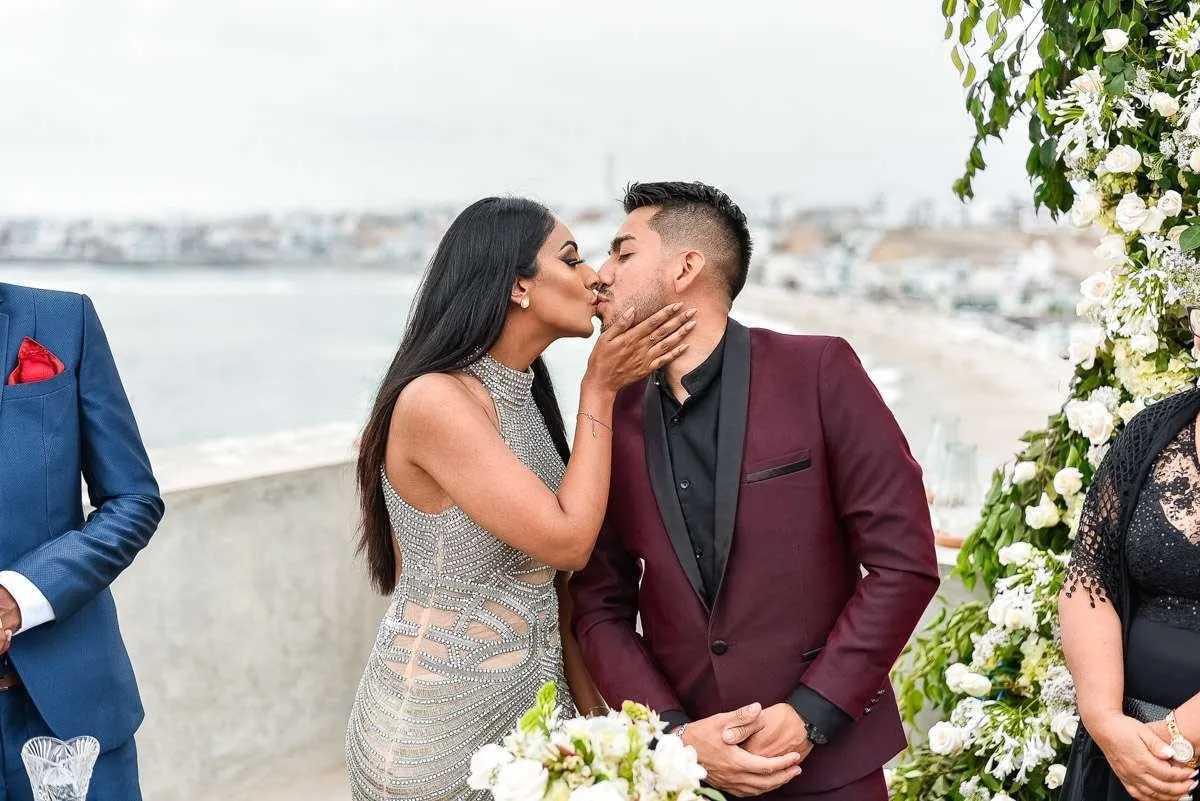 The couple kiss beside an arch of white flowers, with the sea behind them