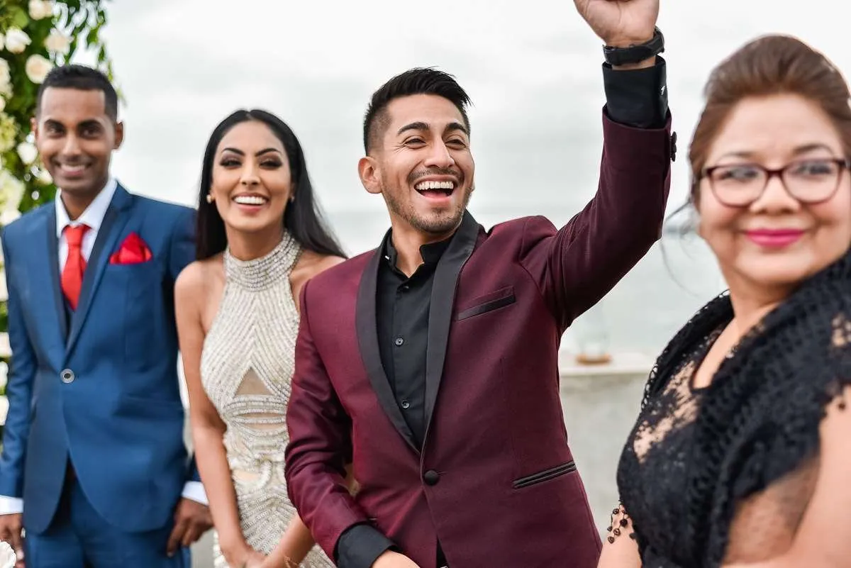 The couple smile beside a guest while a friend raises an arm, laughing