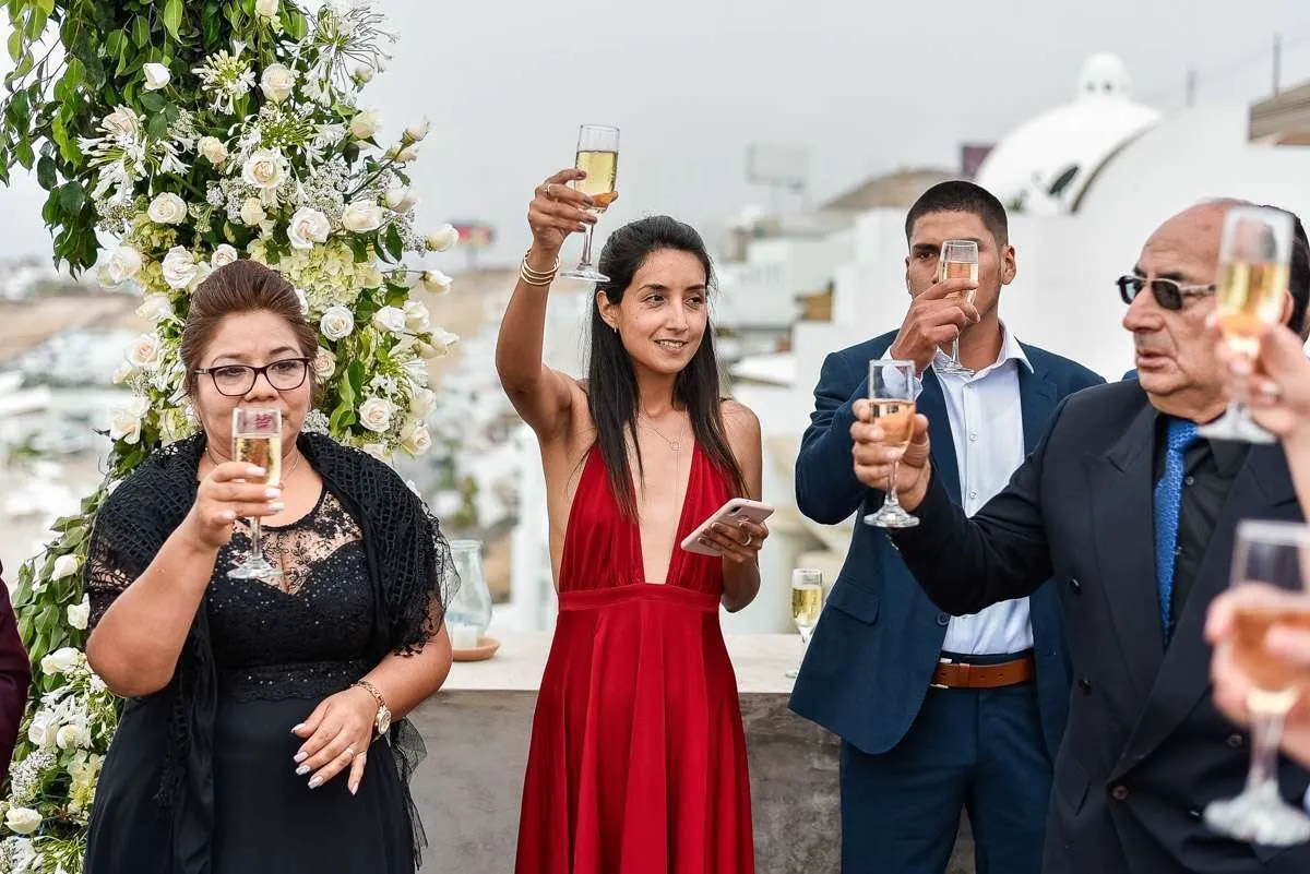 Guests raising a toast beside an arch of white flowers, with white buildings and an overcast sky behind