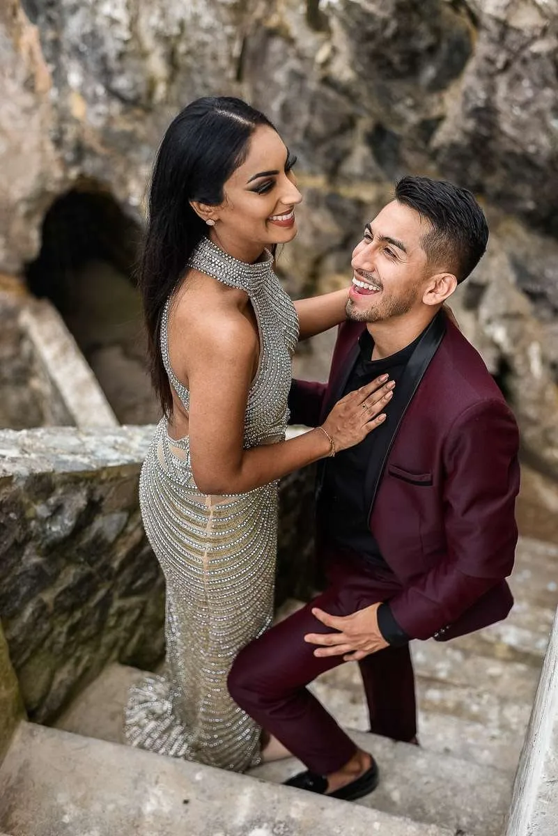 La novia ríe con el novio, que se inclina hacia ella, en una escalera de piedra junto al mar
