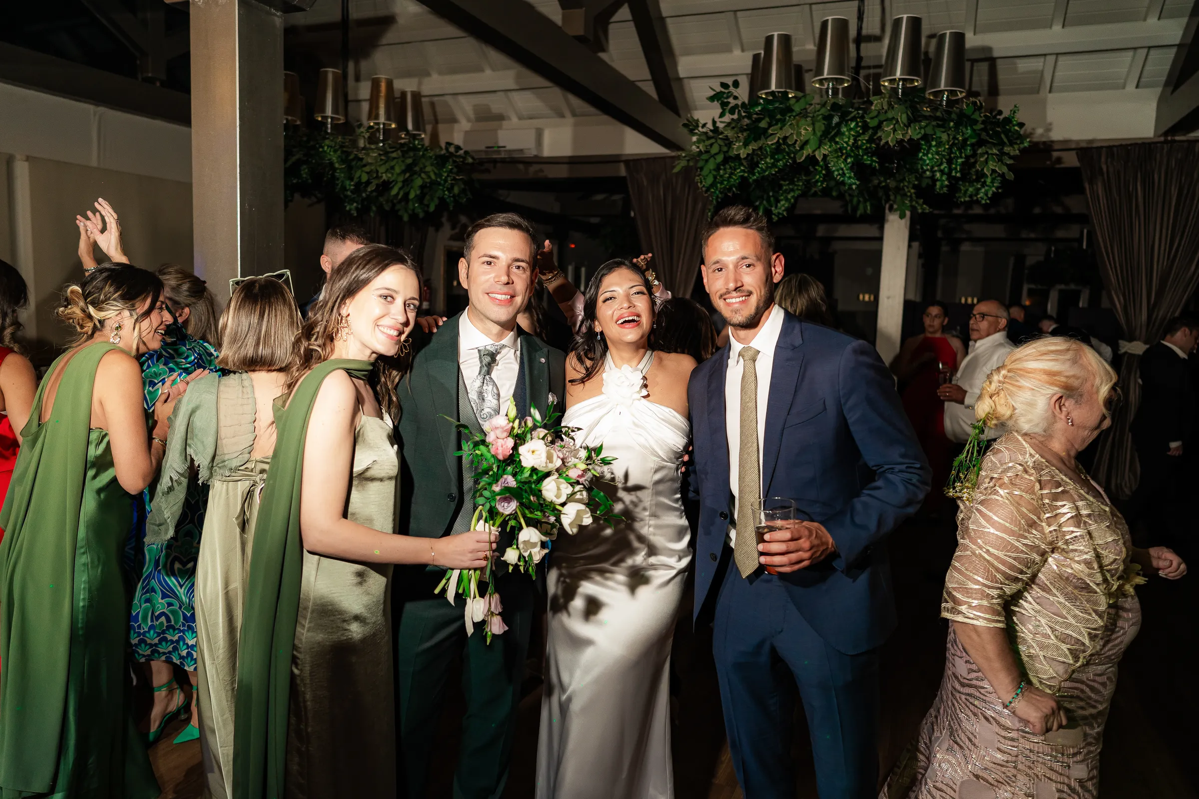A group of guests dressed in green and blue pose beside a woman in white, in a room with wooden beams