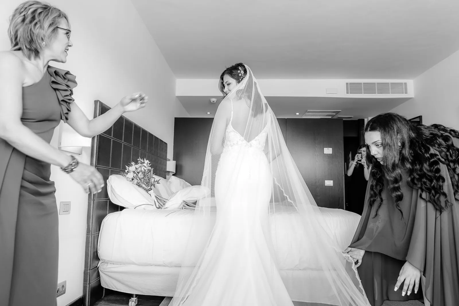 Una novia con velo se prepara en una habitación mientras dos personas la ayudan, en blanco y negro