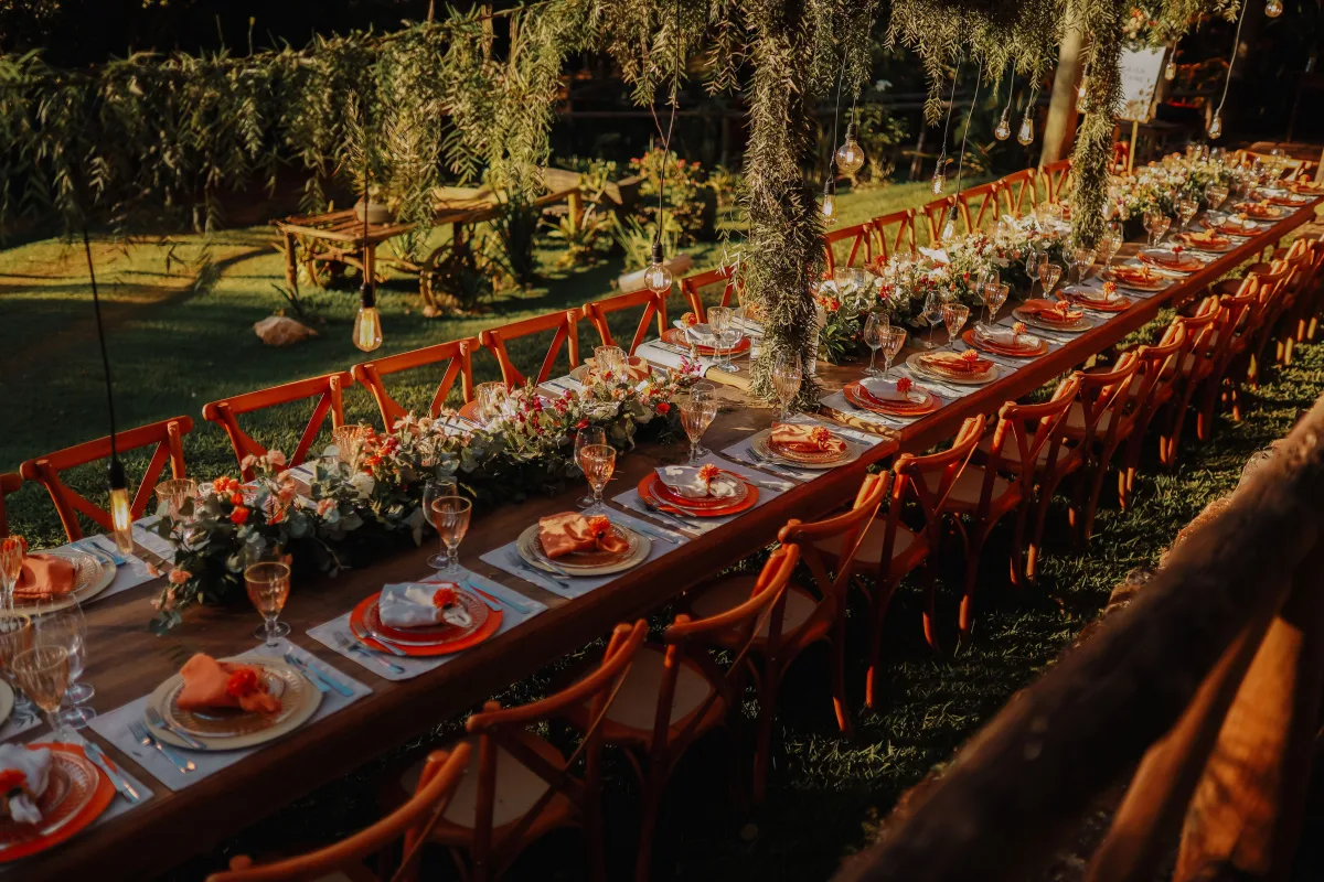 A long wooden table laid for an outdoor dinner, with low flowers, plates and glasses, under a vine canopy with lights