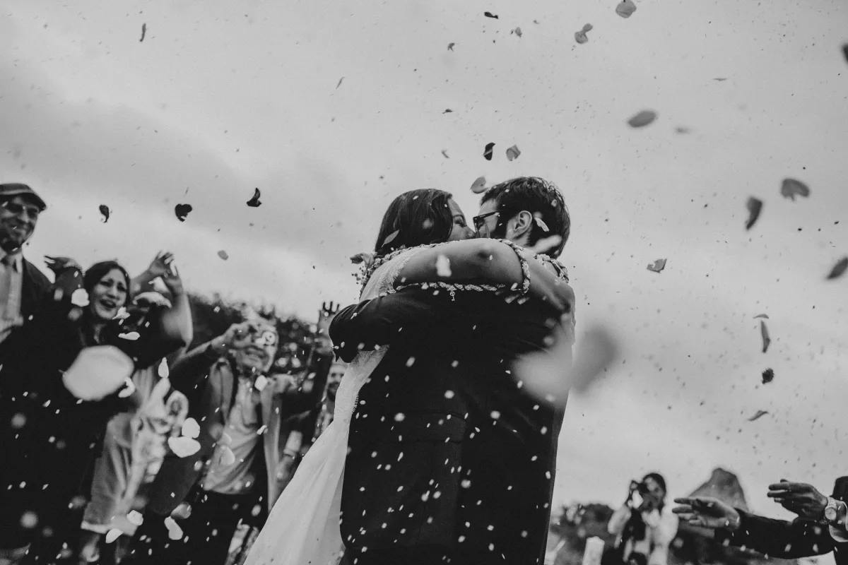 A couple embrace in black and white as guests throw confetti and the pieces fall through the air