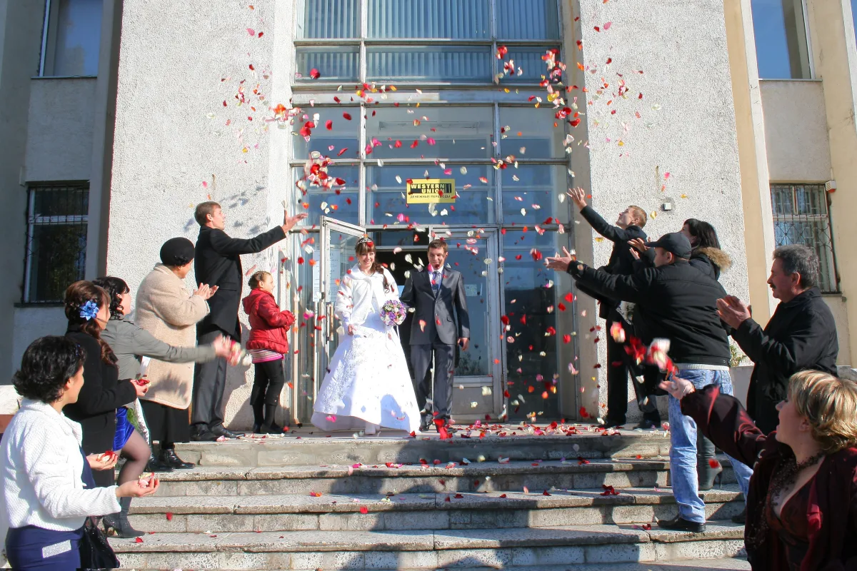 Los novios salen por la puerta de un edificio mientras los invitados, en la escalinata, les lanzan pétalos rojos y rosas