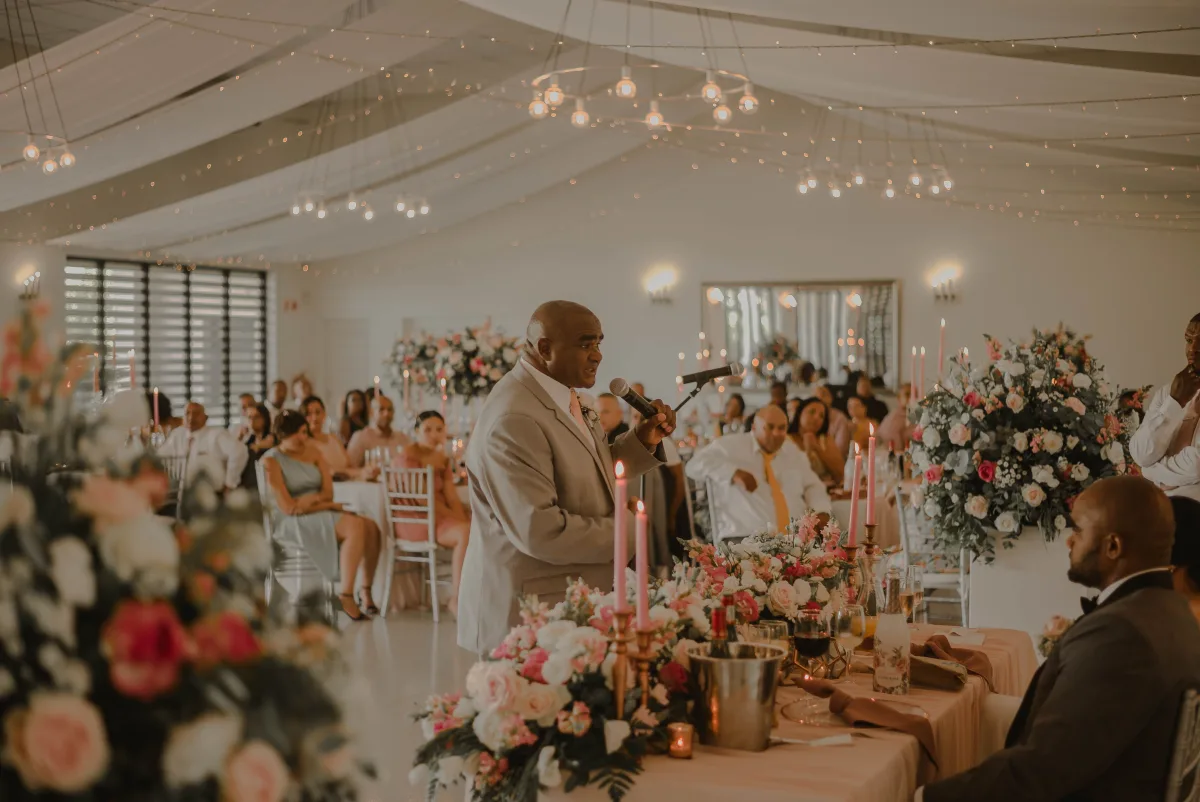 A guest stands to give a speech beside the tables of a bright hall with hanging lights and large flower centrepieces