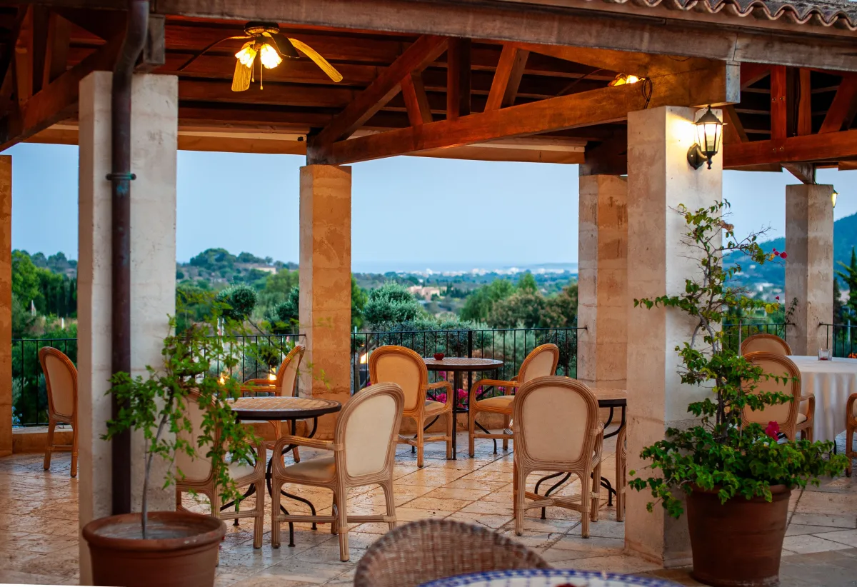 The covered terrace of a hotel, with stone columns, timber beams and tables with wicker chairs facing an open landscape