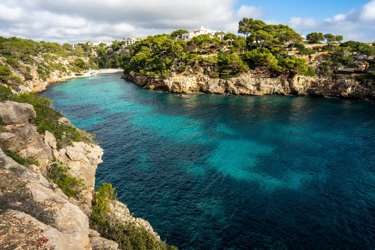 A turquoise cove between cliffs and pines, with white houses on the headland