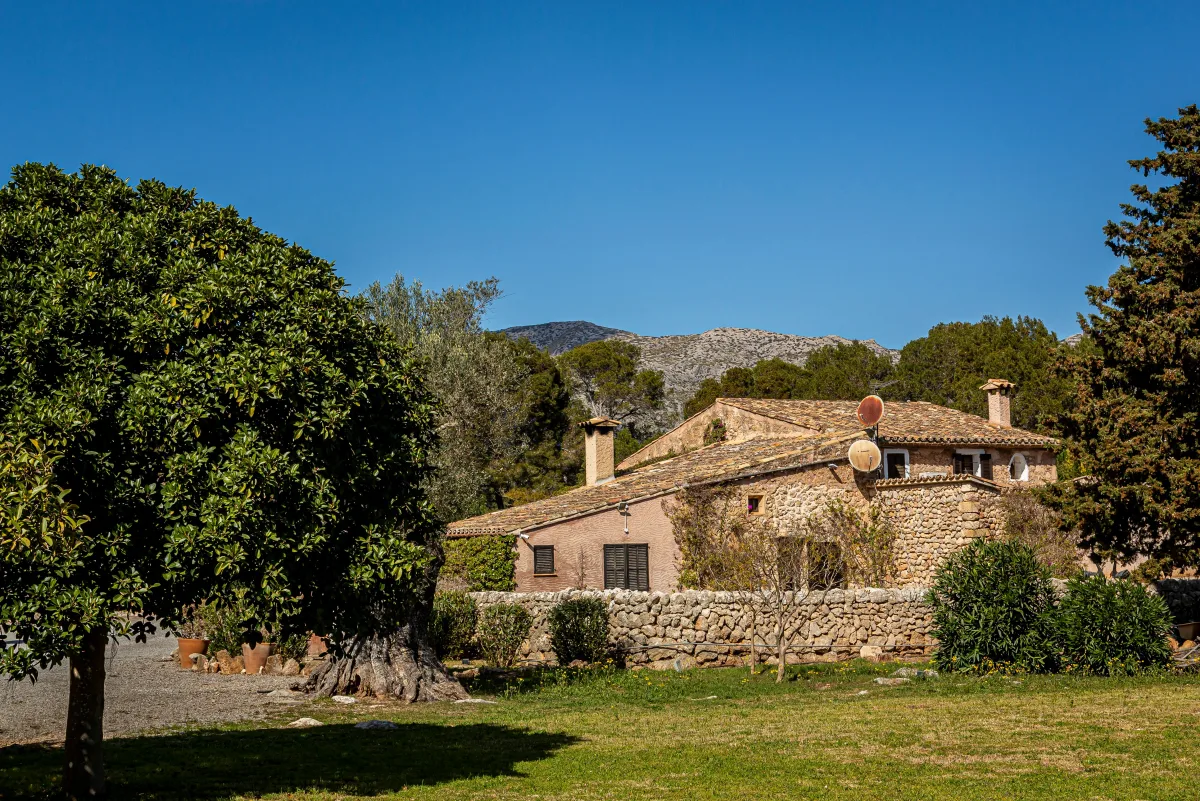 A stone house with a tiled roof in a valley, with orange trees, lawn and a mountain behind