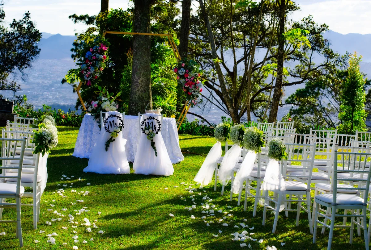 Ceremonia preparada en un jardín: sillas blancas con lazos de tul, un arco floral entre árboles y montañas al fondo