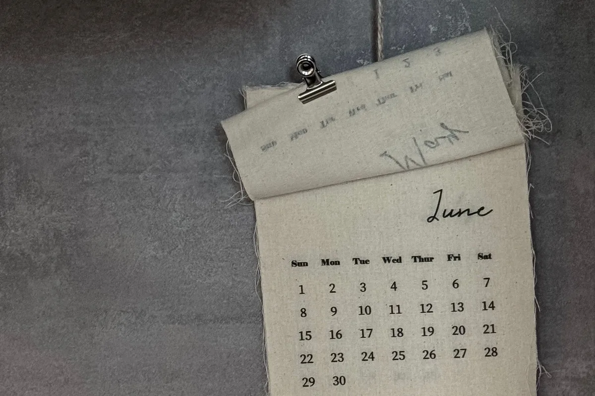 A fabric calendar showing the dates of June, hanging from a clip on a grey wall