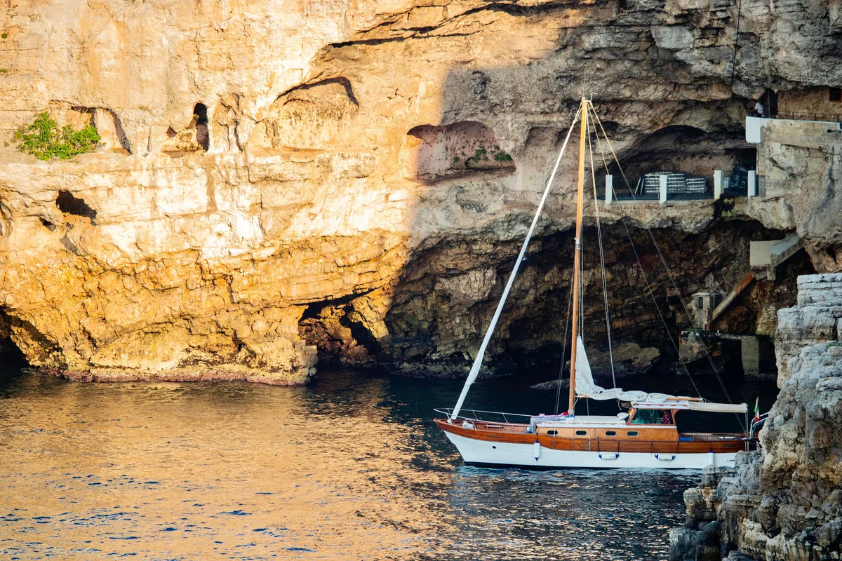 A sailboat moored beside a pale rock cliff, on calm water in warm light