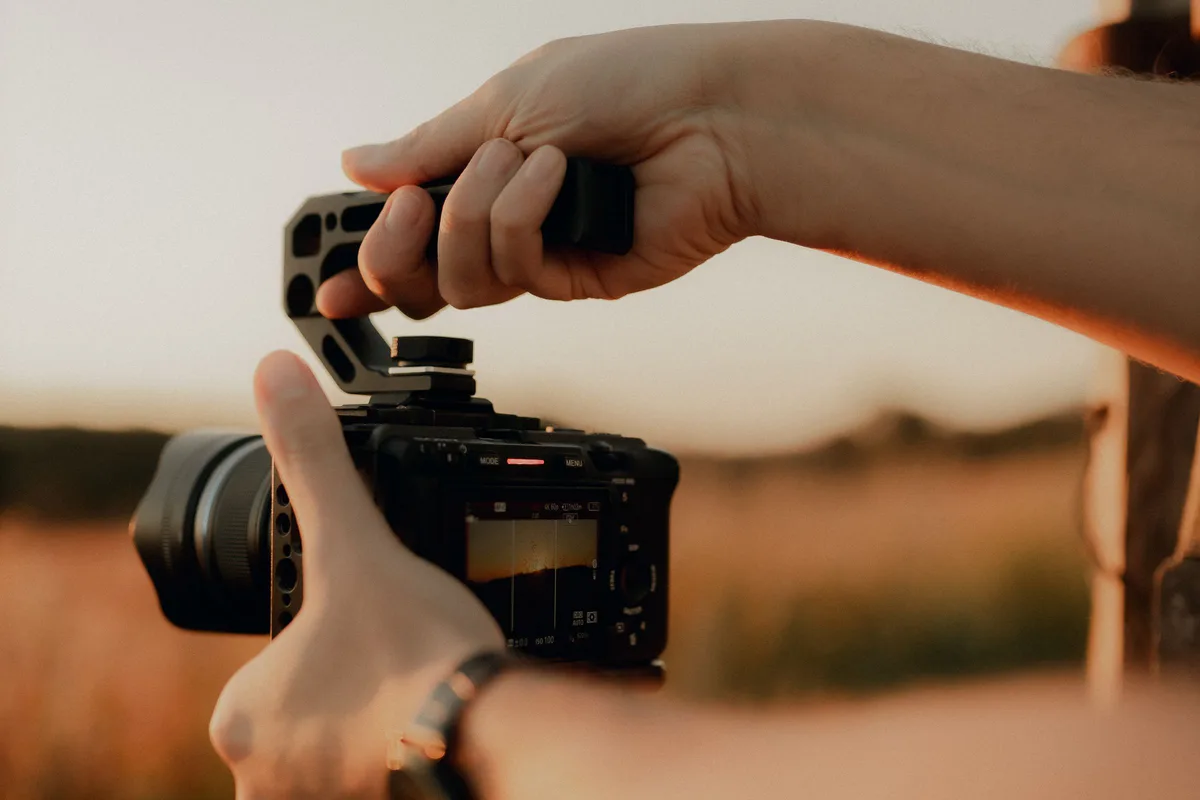 Hands holding a video camera by its handle at sunset, with the field out of focus behind