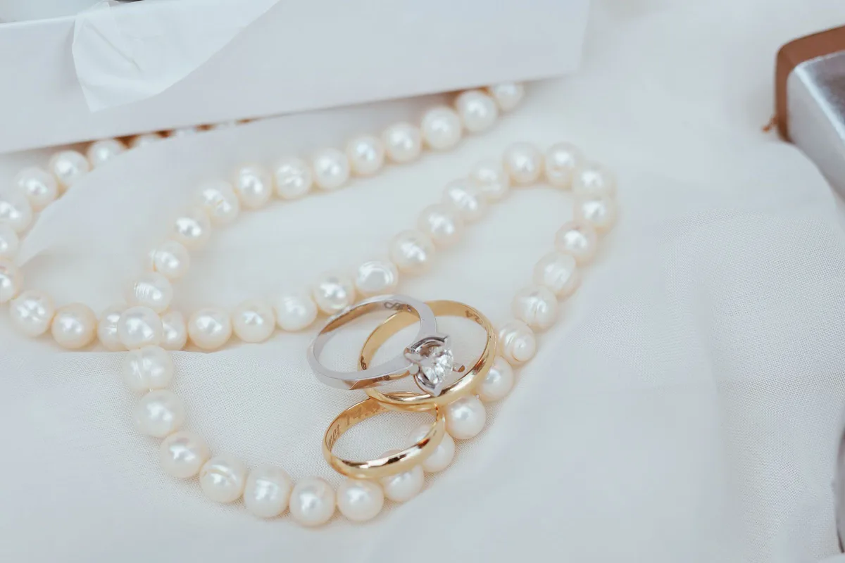 Two gold wedding bands and a stone ring resting on a pearl necklace and white fabric
