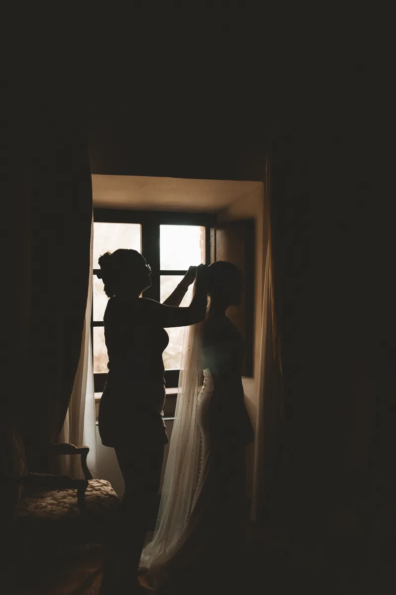 Dos mujeres a contraluz junto a una ventana; una sujeta el velo de la novia
