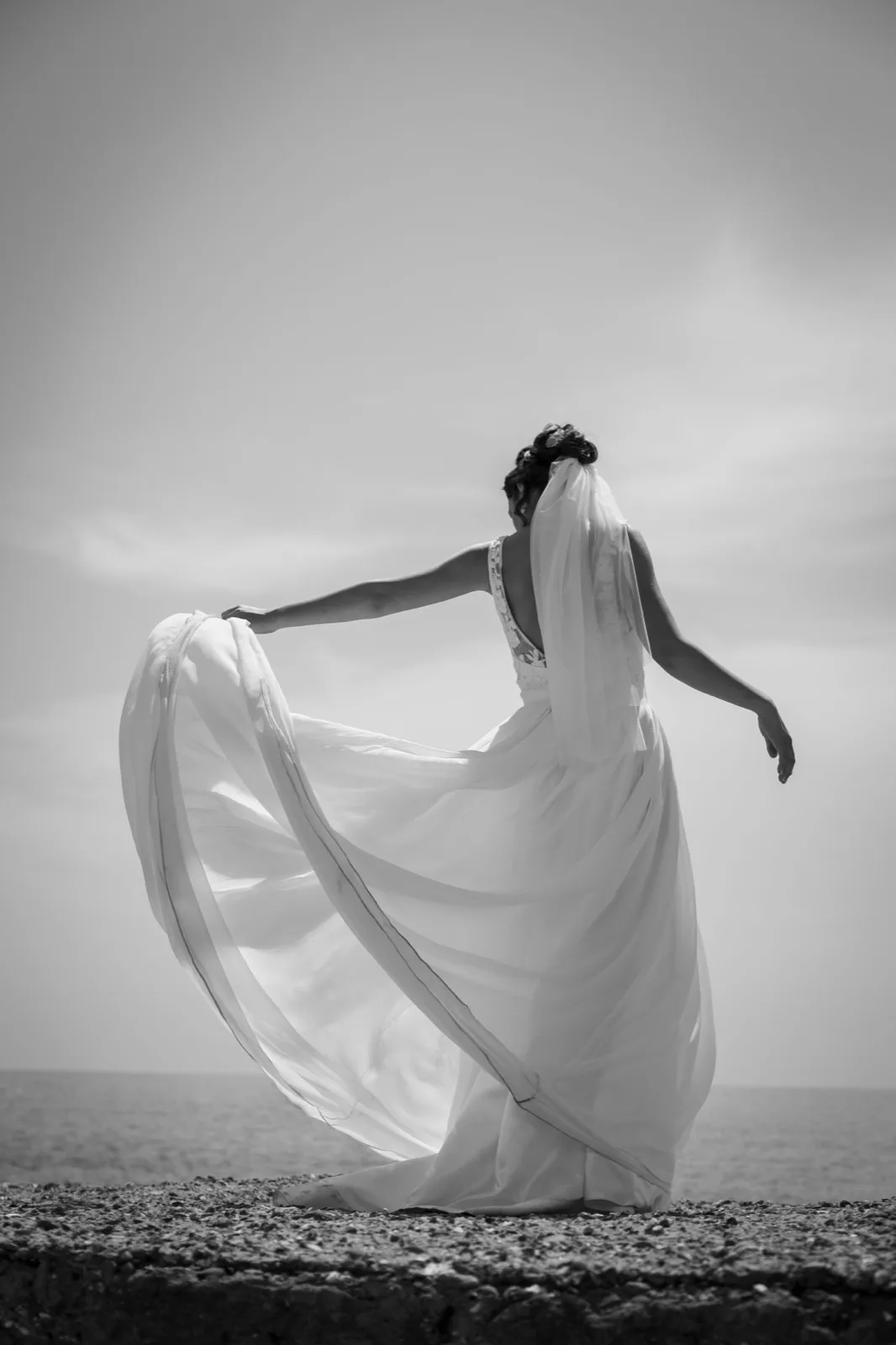 A bride seen from behind, in black and white, her dress and veil moved by the wind before a grey-skied sea