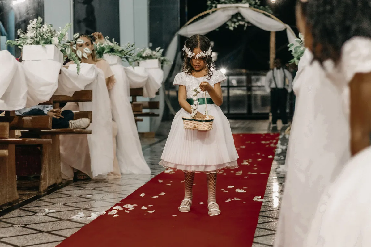 A girl in a flower crown carrying a small basket walks along a red carpet between the pews of a ceremony
