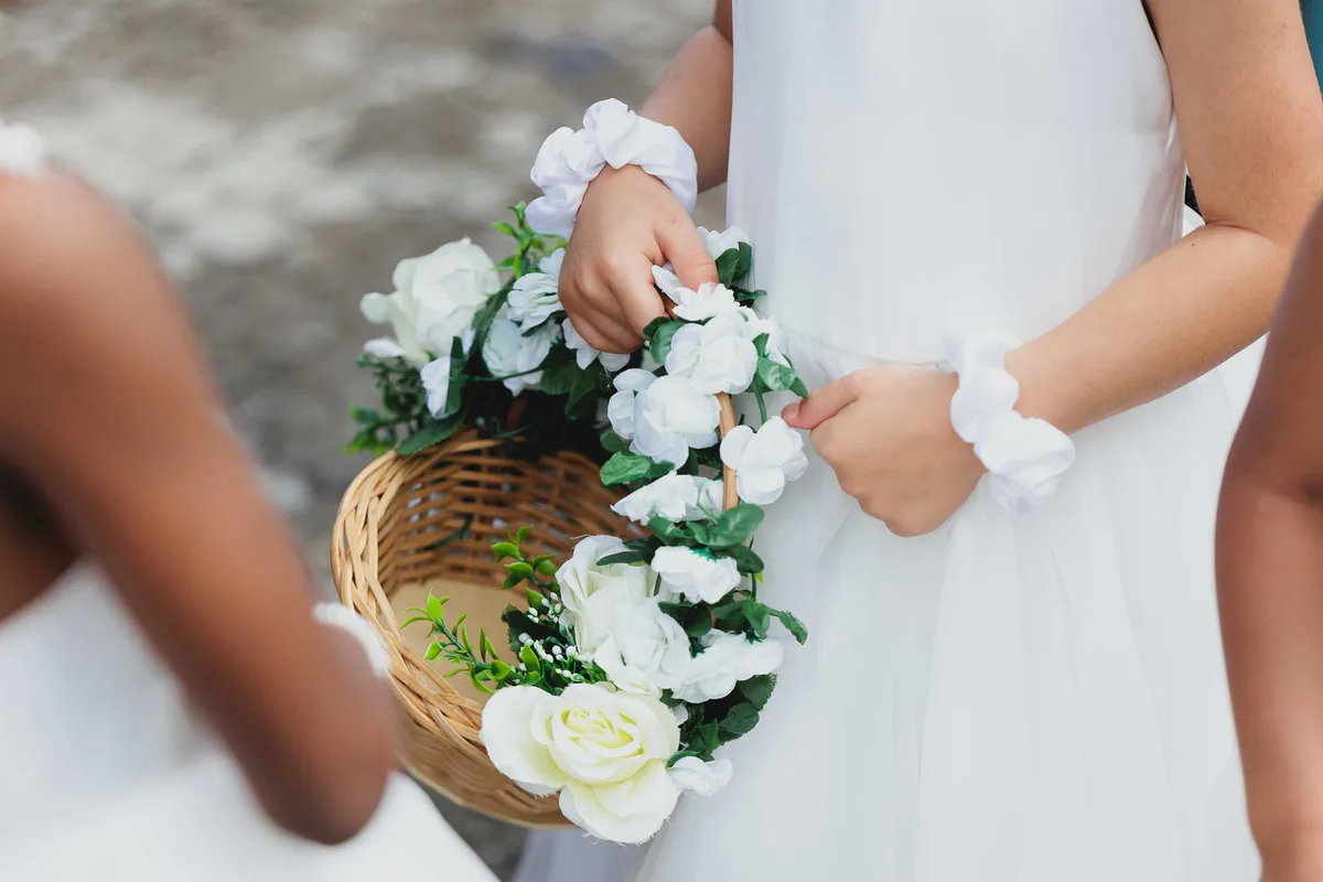 A child holding a basket of white roses and a flower bracelet, beside someone in a white dress