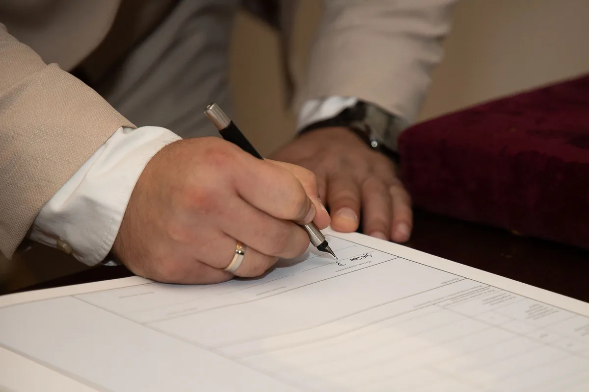 A groom signs a document with a pen, his ring visible on his hand and the paper on the table