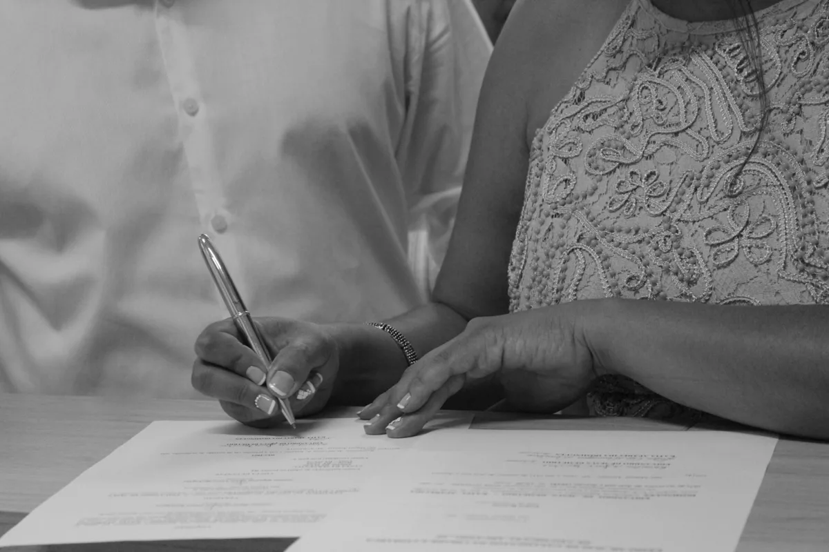 Two people sign a document together, in black and white; only their hands, the pen and the paper are visible