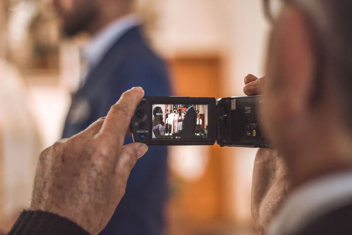 An older person's hands hold a camcorder and film a wedding, with the scene visible on the screen