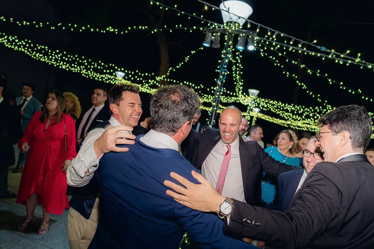 Invitados abrazados y sonrientes, de noche y al aire libre, bajo guirnaldas de luces y un foco colgado