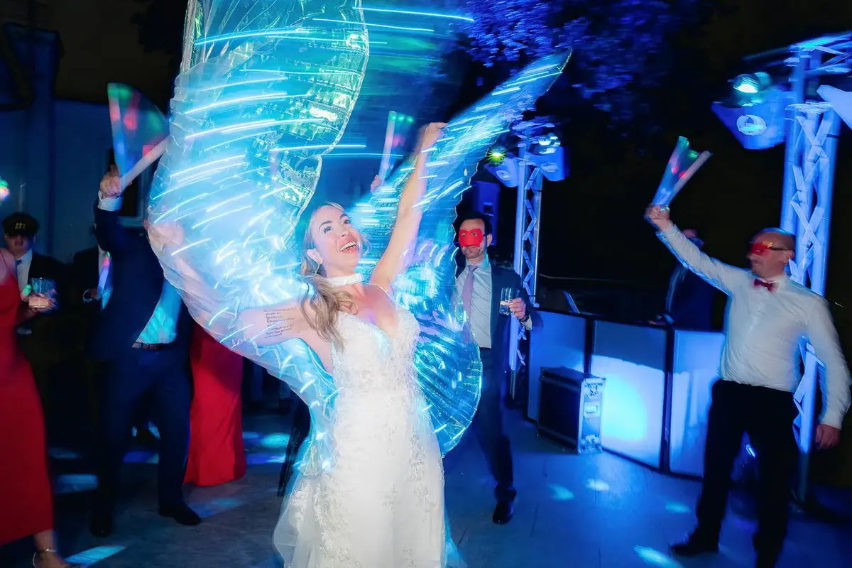 The bride dances with translucent wings that catch the blue light, beside the music table and spotlights, among guests in red masks