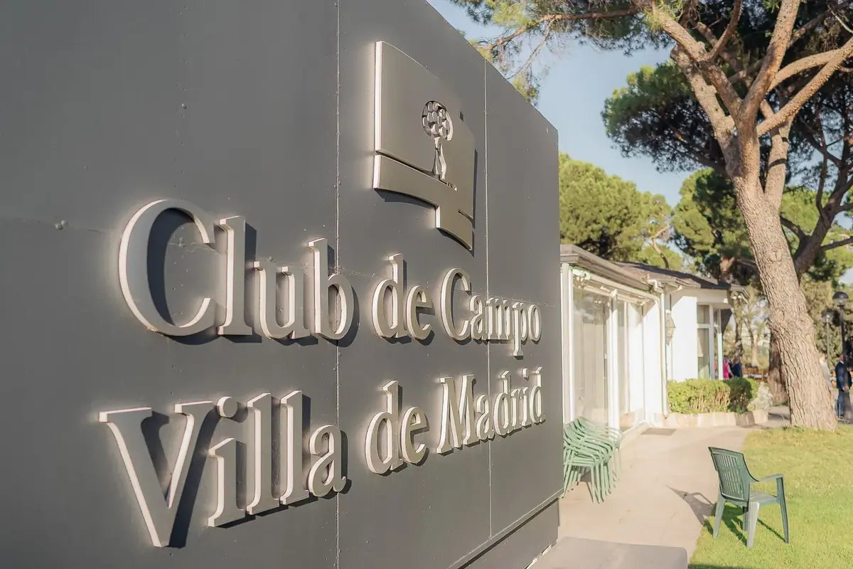 Embossed country club sign among pines, with a garden in daylight