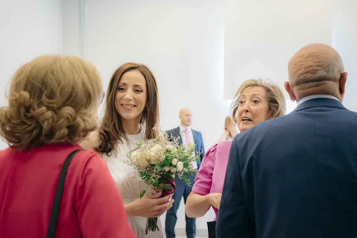 La novia, con un ramo de flores blancas, sonríe mientras conversa con varias personas que la rodean en una sala luminosa, dos de ellas de espaldas