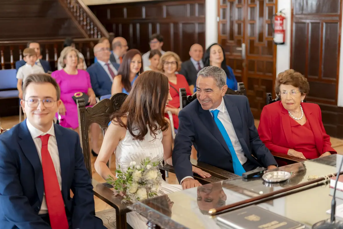 The couple seated with their relatives at a table, in a room with dark wood panelling; the groom smiles in a blue suit and red tie