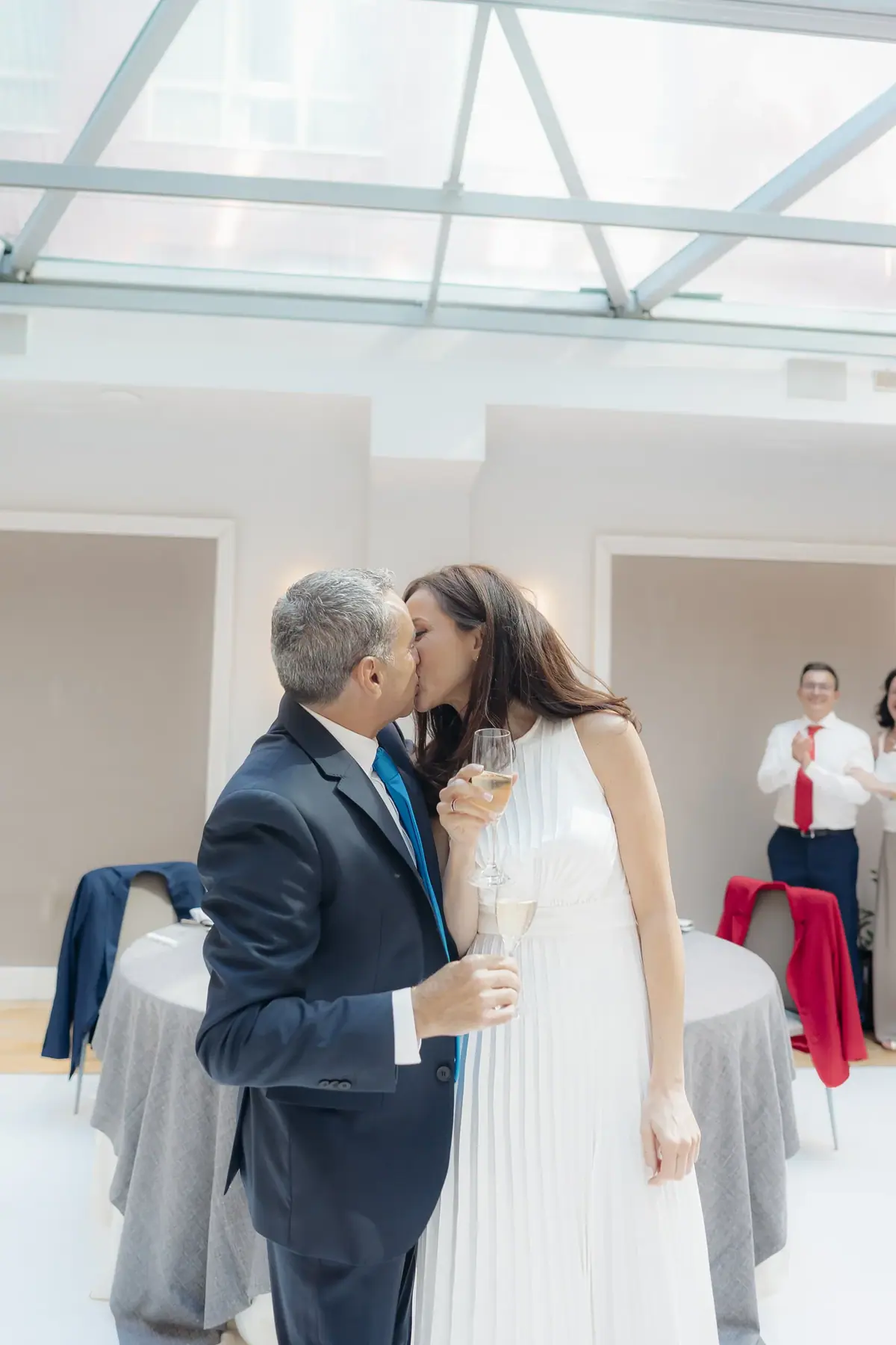 La novia, de blanco y con una copa en la mano, besa a su pareja en un salón con techo de cristal mientras al fondo un invitado aplaude